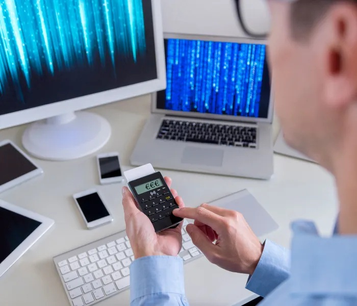 Person using a smartphone with a financial app. A computer and laptop display digital code. Multiple devices are on the desk.