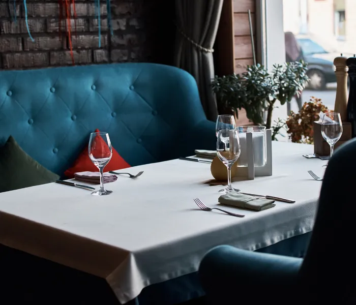 Table set for four with wine glasses, napkins, and cutlery in a restaurant with blue seating and a window view.