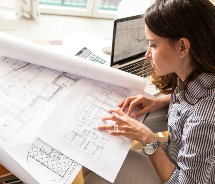 A woman reviews architectural blueprints at a desk with a laptop and calculator.