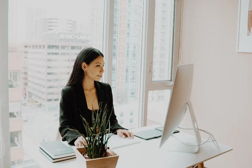 A person sits at a desk in an office, working on a computer. There's a plant and notebooks on the desk, with large windows behind.