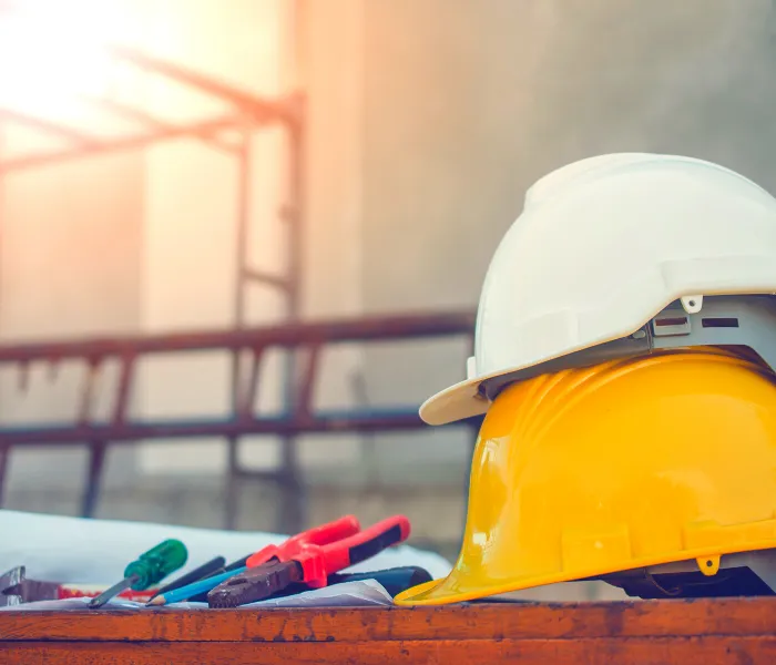 Two hard hats, one white and one yellow, sit on a table with various tools and rolled blueprints.