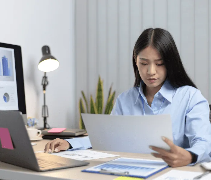 A woman in a blue shirt reviews documents at a desk with a laptop, lamp, and charts displayed on a computer monitor.